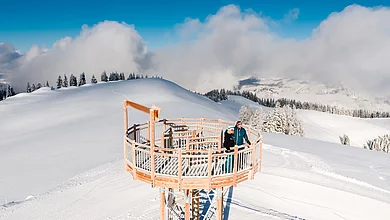 Ein Paar auf einem Aussichtsturm auf einem Berg schaut sich die Winterlandschaft des Saalachtals an